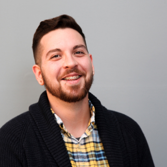 Tyler, on a gray background, facing the camera and smiling in a headshot taken at Midwest UX 2018. He is wearing a yellow, black, and blue plaid button-up with a dark blue, waffle-knit cardigan on top.