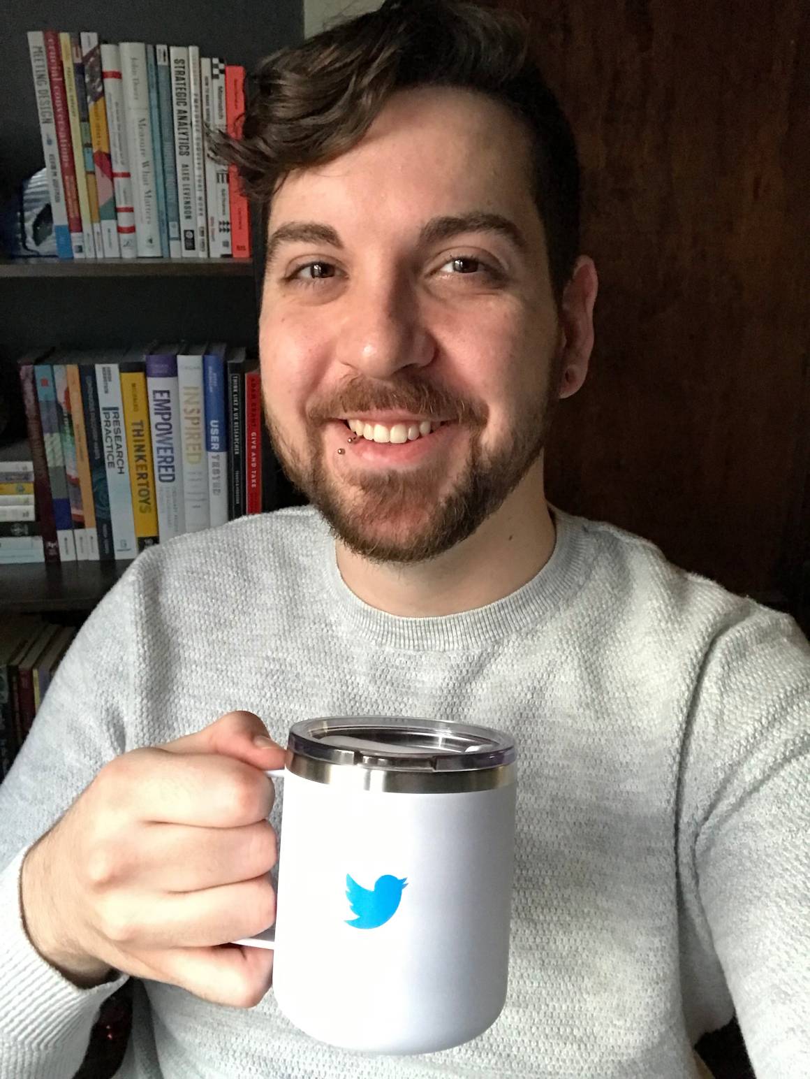 Tyler wearing an off-white sweater and smiling while holding his Twitter mug, in front of a bookshelf.